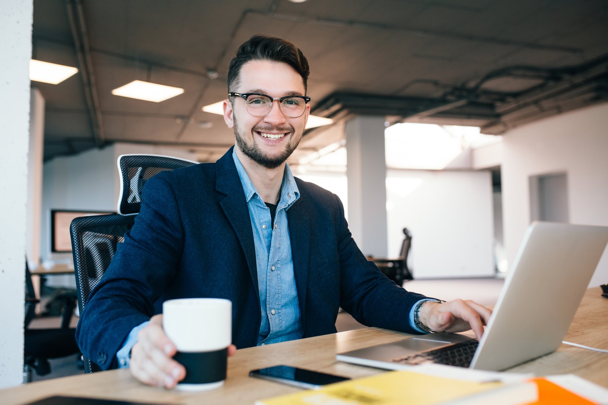 atractivo-hombre-de-pelo-oscuro-esta-trabajando-en-la-mesa-en-la-oficina-viste-camisa-azul-con-chaqueta-negra-esta-tomando-una-taza-de-cafe-y-sonriendo-la-camara atractivo-hombre-de-pelo-oscuro-esta-trabajando-en-la-mesa-en-la-oficina-viste-camisa-azul-con-chaqueta-negra-esta-tomando-una-taza-de-cafe-y-sonriendo-la-camara